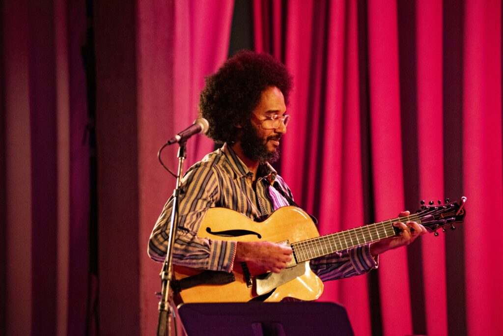 Photo of guitarist Geovane Santos wearing a striped shirt sitting in front of a red curtain and holding a guitar.