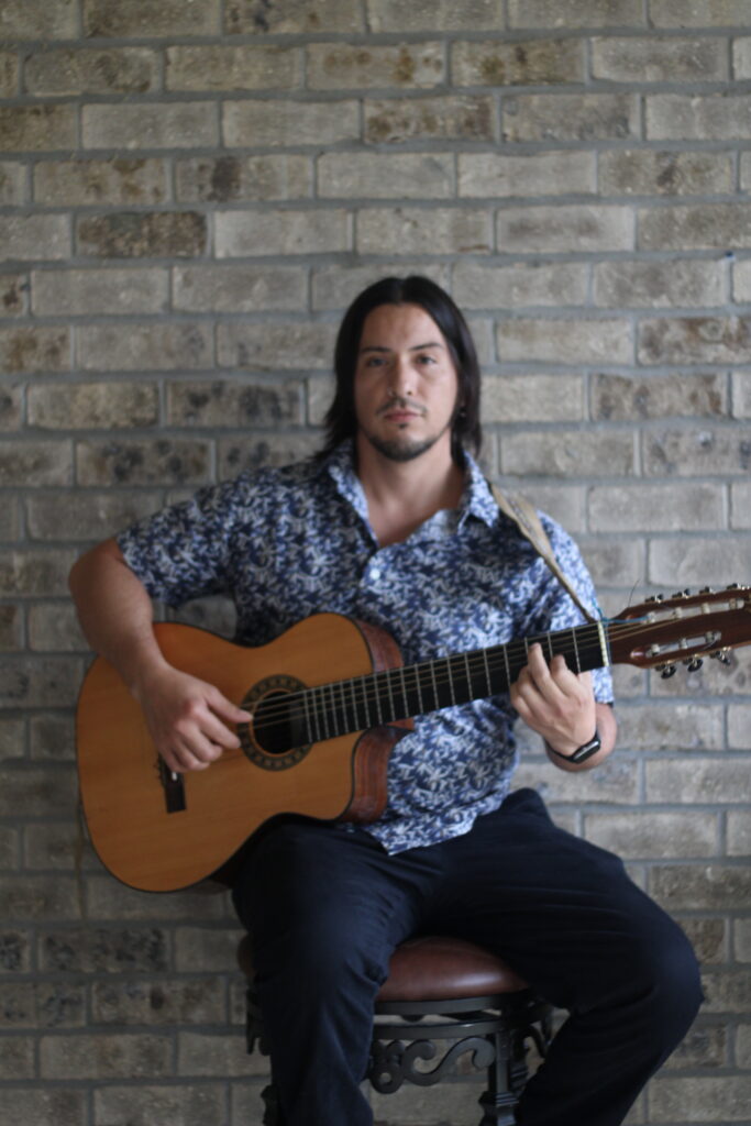 A man holding a guitar sitting in front of a brick wall